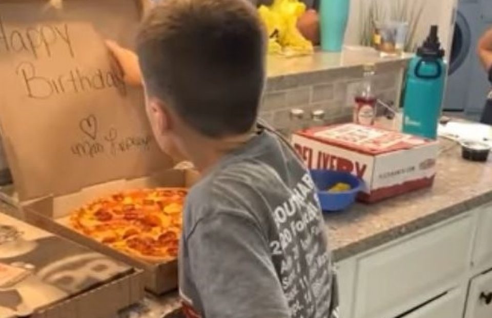 A boy in a gray T-shirt watches as an adult in a baseball cap prepares to cut a pizza in a kitchen decorated with a bunny wall hanging. The pizza box is open on the counter.
