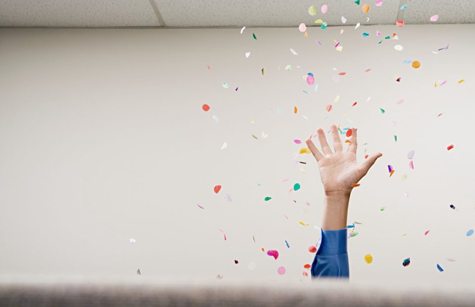 A person's hand and arm raised, wearing a blue uniform sleeve, reaching up amidst colorful confetti falling in a celebration scene.