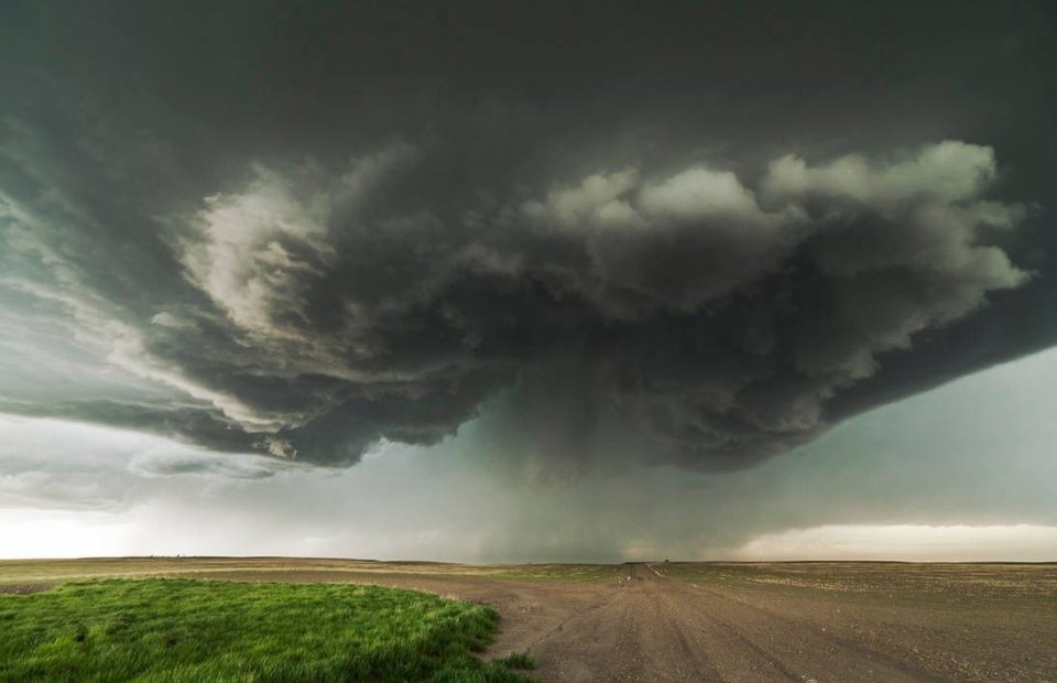 Dark storm cloud with a possible tornado forming over a flat rural landscape, with green grass and a dirt road leading into the distance.