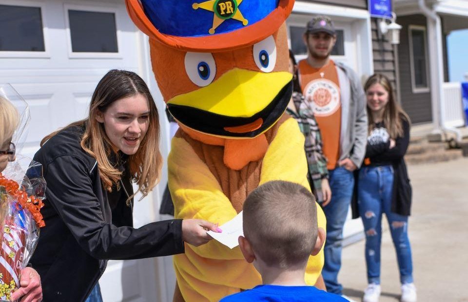A person dressed in a duck mascot costume with a police badge hat interacts with a young boy, handing him a piece of paper, outside a house on a bright day.
