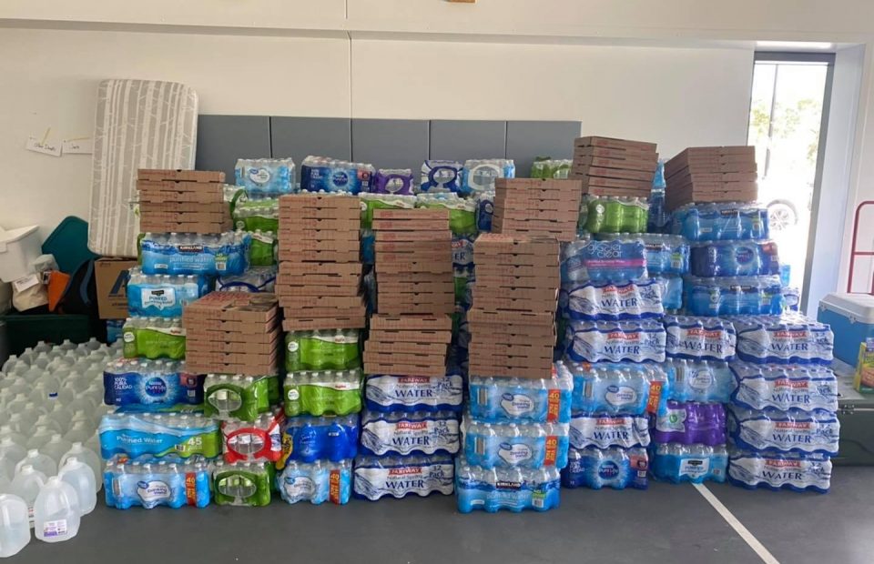 Stacks of bottled water and pizza boxes arranged on a table in a bright room with a window in the background. Two people are partially visible behind the items.