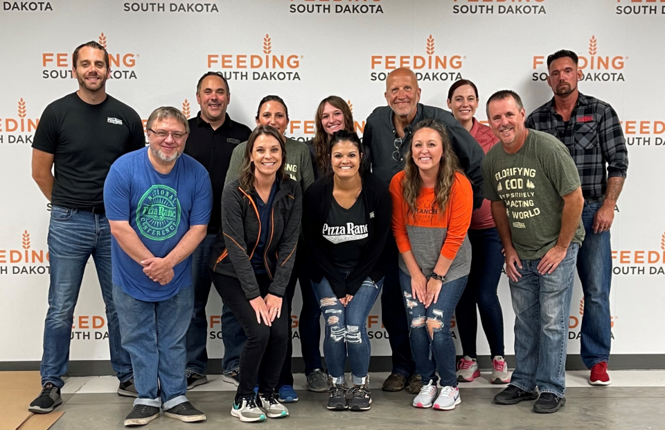 Group of people smiling and posing in front of a "Feeding South Dakota" backdrop, with some wearing Pizza Ranch attire, in an indoor setting.