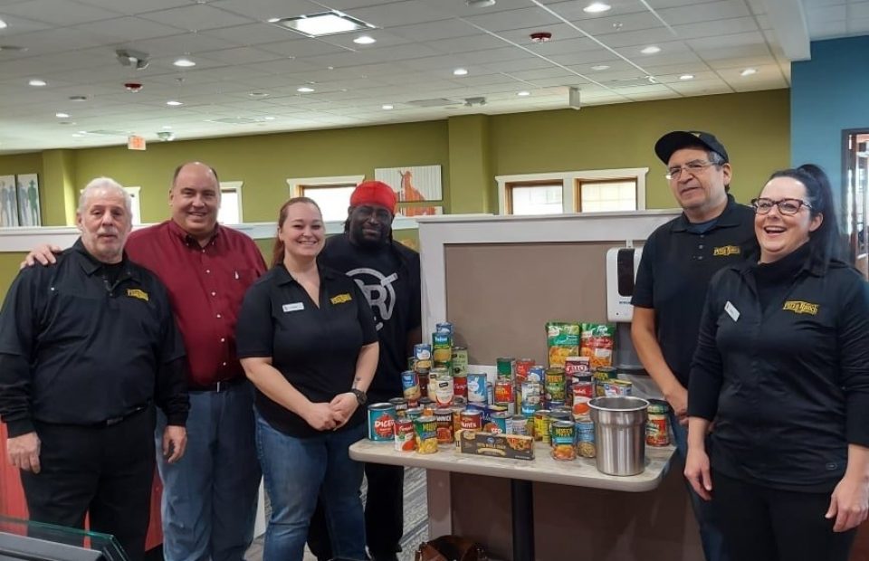 Six people standing in a restaurant interior, five wearing Pizza Ranch uniforms and one in a red shirt, smiling behind a table with assorted canned and boxed food items.
