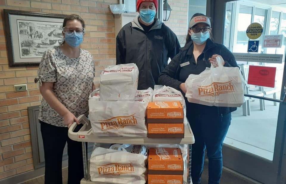 Three employees wearing masks and Pizza Ranch uniforms stand behind a cart filled with takeout pizza boxes and bags inside a restaurant.