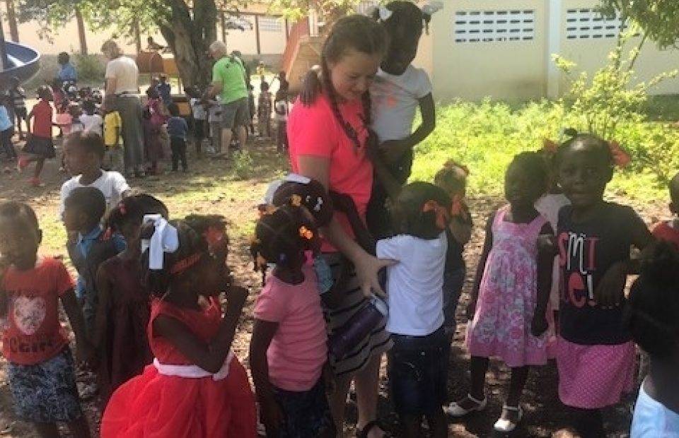 Children gathered outdoors under trees, some dressed in colorful clothing, with a young woman interacting with a girl in a pink shirt.