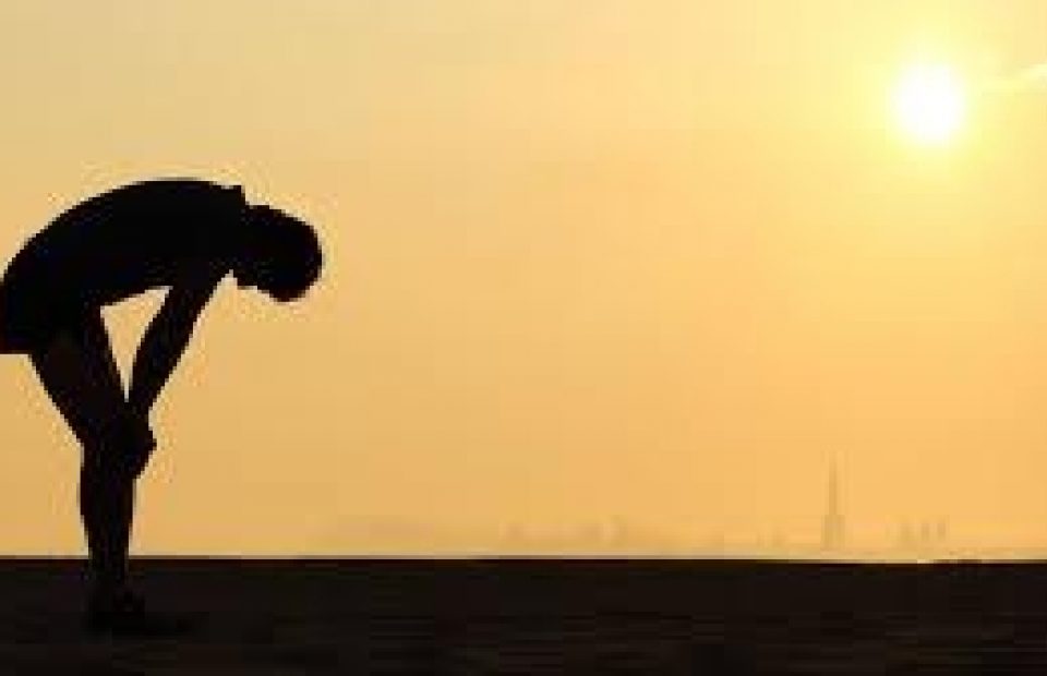A person bending forward at sunset, appearing to be stretching or exhausted, with a bright sun low on the horizon and calm sky in the background.