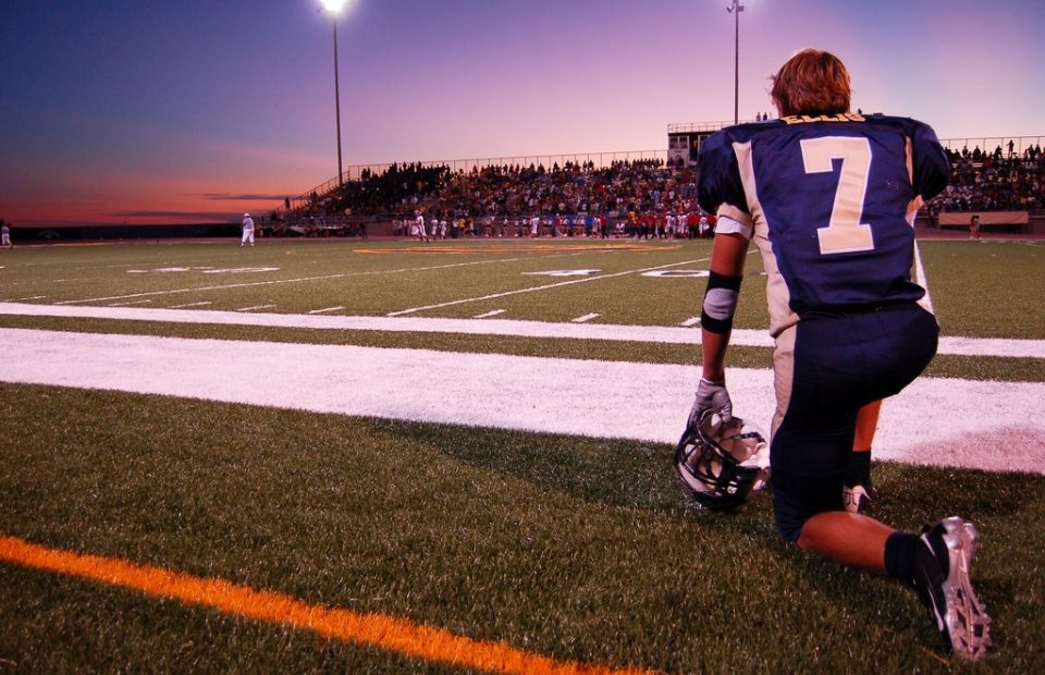A football player kneeling on the field during dusk, with a large crowd in the stands and bright stadium lights illuminating the scene.