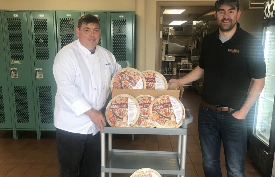 Two men, one in a white chef's coat and the other in a black Pizza Ranch uniform, stand beside a cart with packaged frozen pizzas in a kitchen or locker room.