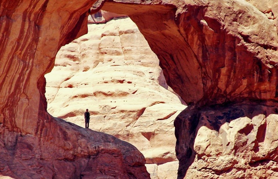 A person stands beneath a natural rock arch in a desert landscape with layered, reddish sandstone formations and a clear, bright sky.