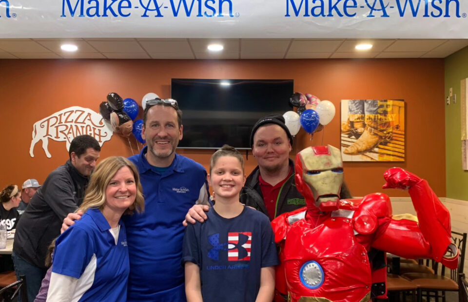 A group of four people and a person in an Iron Man costume pose together inside a pizza restaurant with a "Make-A-Wish" banner overhead.