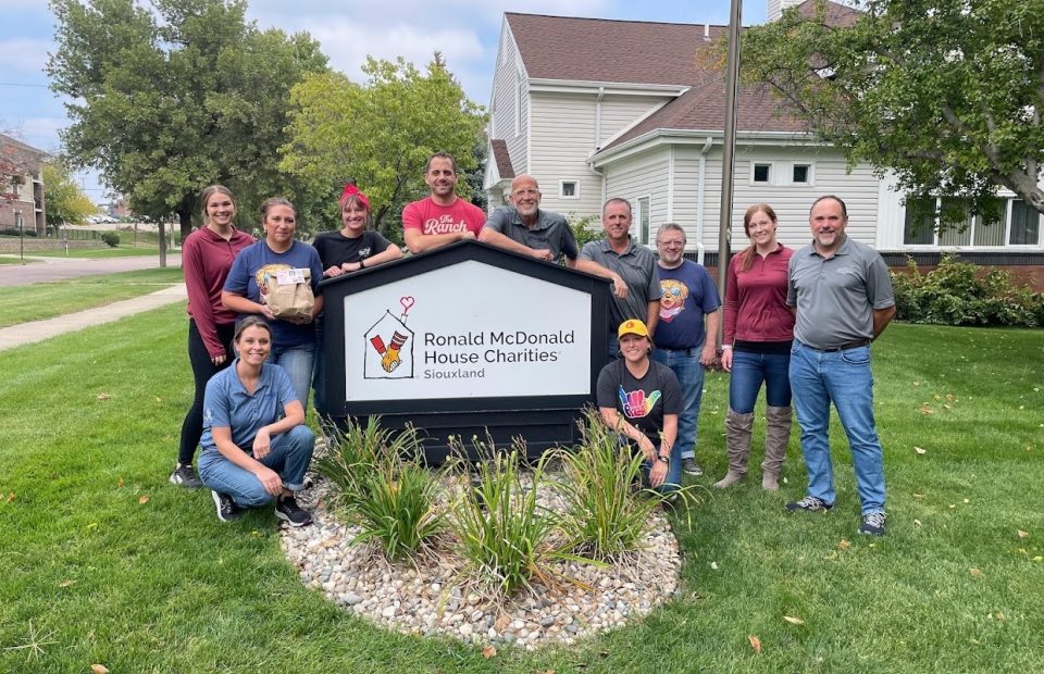 A group of ten people poses outdoors around a sign for Ronald McDonald House Charities in Siouxland, with trees and a residential building in the background.