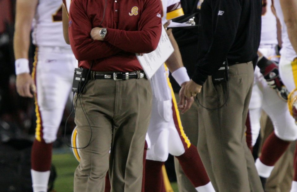 A football coach wearing a maroon cap and long-sleeve shirt with a headset, standing with arms crossed on the sideline of a game, surrounded by players in white and maroon uniforms.