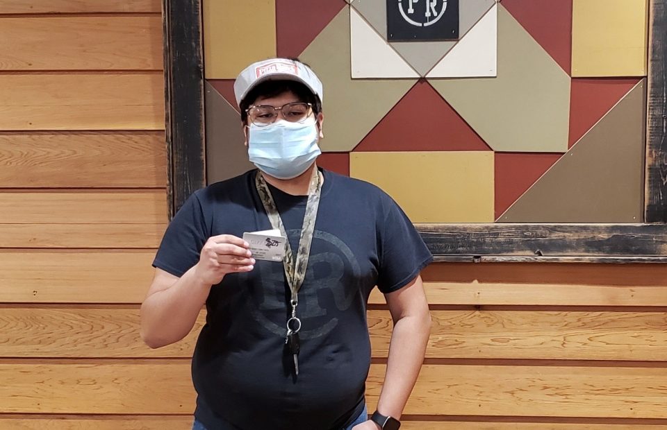 A person wearing a face mask, glasses, and a cap holding a card, standing in front of a wooden wall with a geometric sign that says "PR".