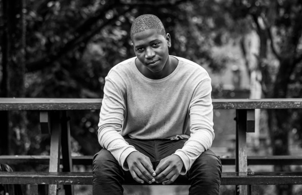 A young man is sitting on a wooden park bench outdoors, wearing a long-sleeved shirt and dark pants, with trees and blurred background in black and white.