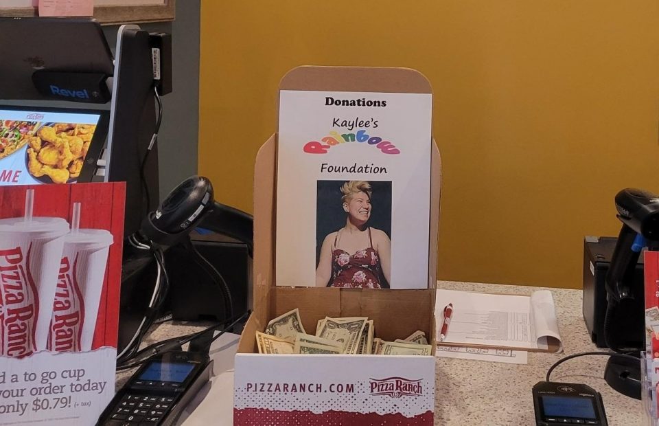 A donation box labeled "Kaylee's Rainbow Foundation" with cash inside. The counter displays menu flyers and POS devices, all branded with Pizza Ranch.