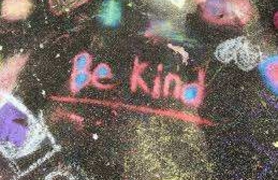 A chalkboard with "Be Kind" written in pink and red with a small heart shape, surrounded by colorful chalk smudges in a casual, creative setting.