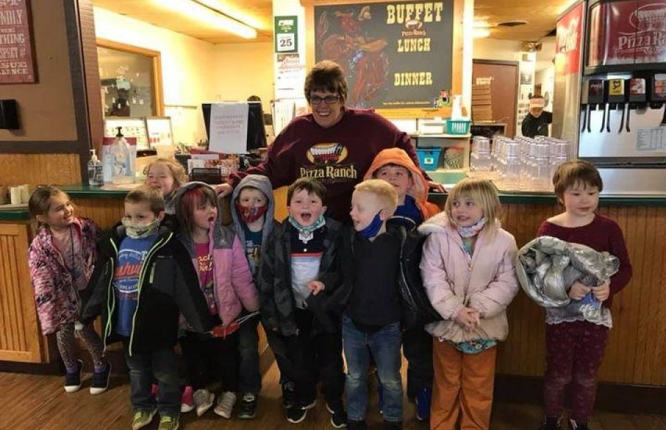 A group of young children and an adult with glasses, all smiling, standing together inside a restaurant with a "Buffet Lunch Dinner" sign behind them.