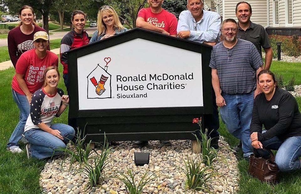 A group of nine people, some wearing Pizza Ranch shirts, pose outdoors around a sign for Ronald McDonald House Charities Siouxland, with trees and houses in the background.