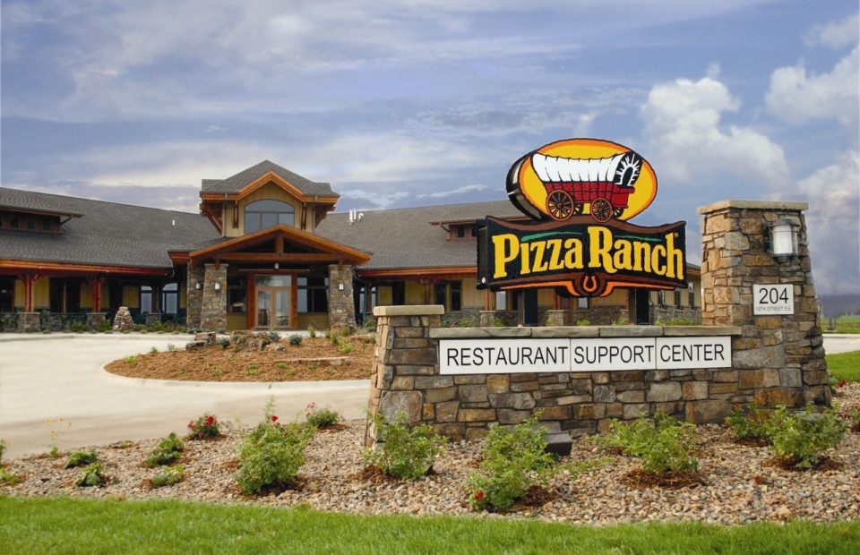 A stone sign with a pizza wagon logo and "Pizza Ranch" text, located in front of a restaurant building with a landscaped foreground and a cloudy sky.