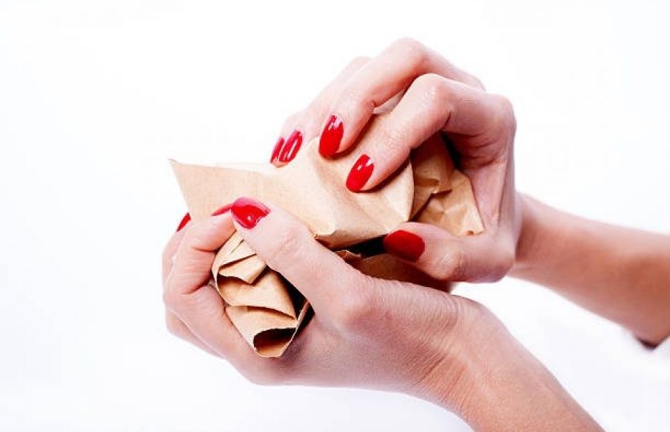 Person with red painted nails squeezing a crumpled brown paper bag against a white background.