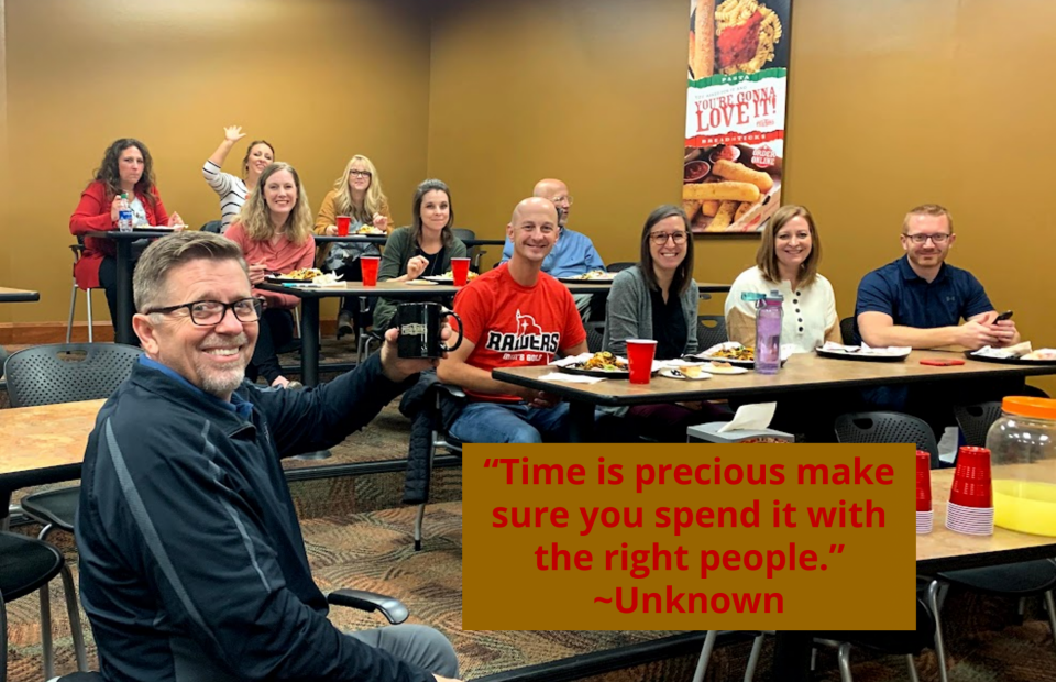 Group of nine adults sitting at tables in a room, smiling and enjoying a meal; a man in the front waves, holding a mug. A quote on a brown box reads, “Time is precious make sure you spend it with the right people.”