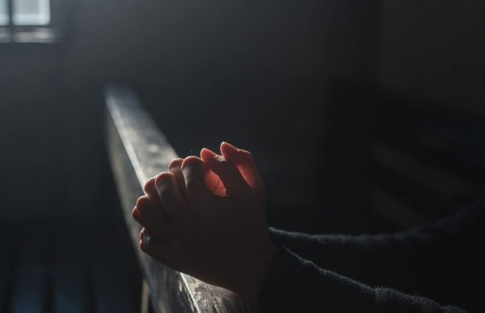 Clasped hands resting on a railing in a dimly lit setting, with subtle sunlight highlighting the fingers and part of the arm.