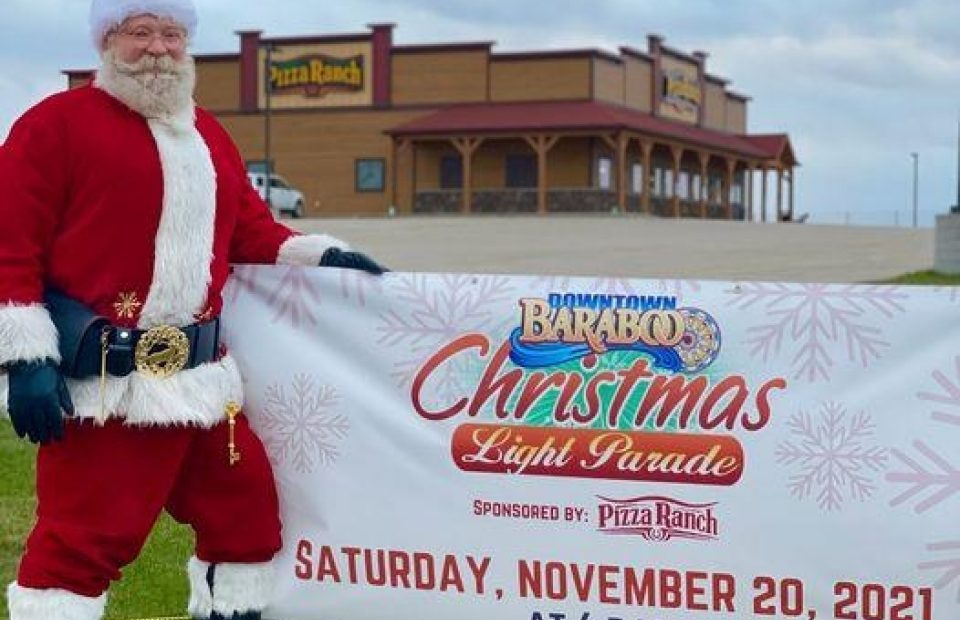 A man dressed as Santa Claus in a red suit with white trim, black boots, and a Santa hat, standing outside on grass, holding a large sign for a Christmas light parade. The background features a Pizza Ranch restaurant.
