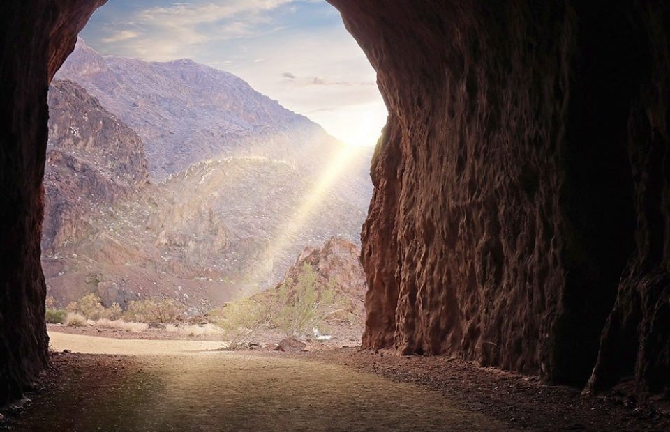 Sunlight streams into a rugged mountain cave, illuminating the rocky cliffs and desert landscape outside.
