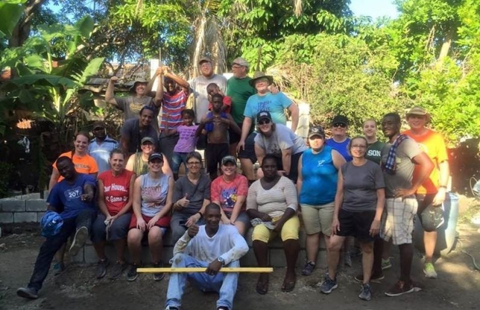 Group of diverse people outdoors in a tropical garden, some sitting, some standing, with trees and lush greenery in the background.