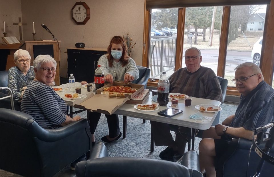 A group of five people, mostly elderly, are gathered around a table with pizza, soda, and snacks in a cozy room with large windows showing parked cars outside.