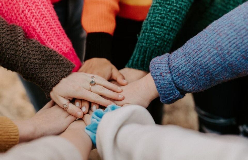 Multiple hands stacked together, some with rings, wearing colorful sweaters, signifying unity and teamwork. Some hands overlap, with a blurred background.