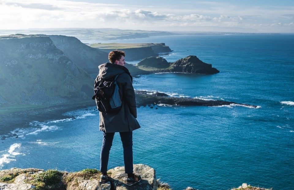 A person standing on a rocky ledge overlooking a rugged coastline with cliffs, green fields, and the ocean under a partly cloudy sky.