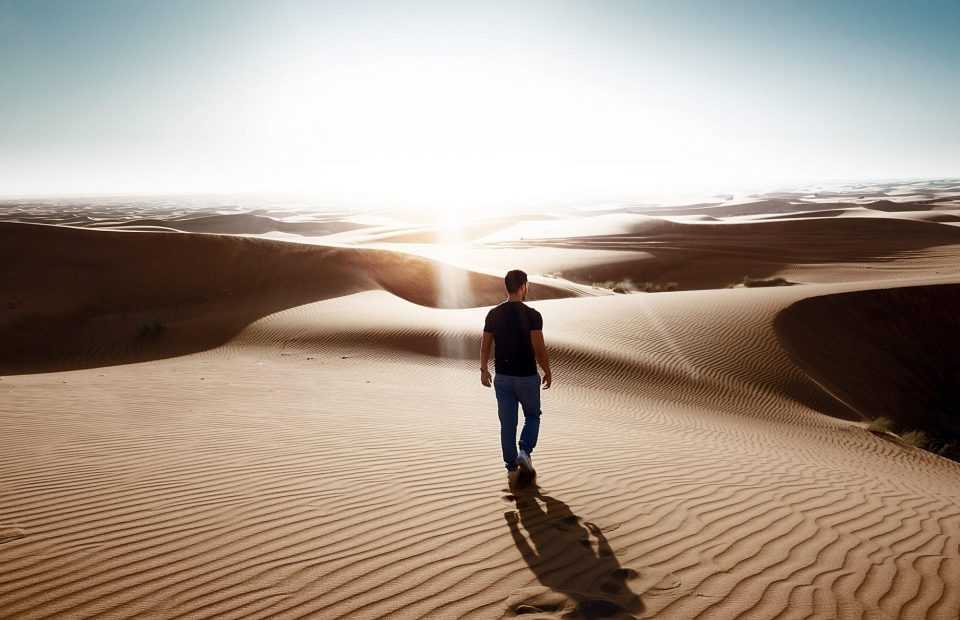 A man in a black t-shirt and blue jeans walking alone across sand dunes at sunset, casting a long shadow over the rippled desert surface.