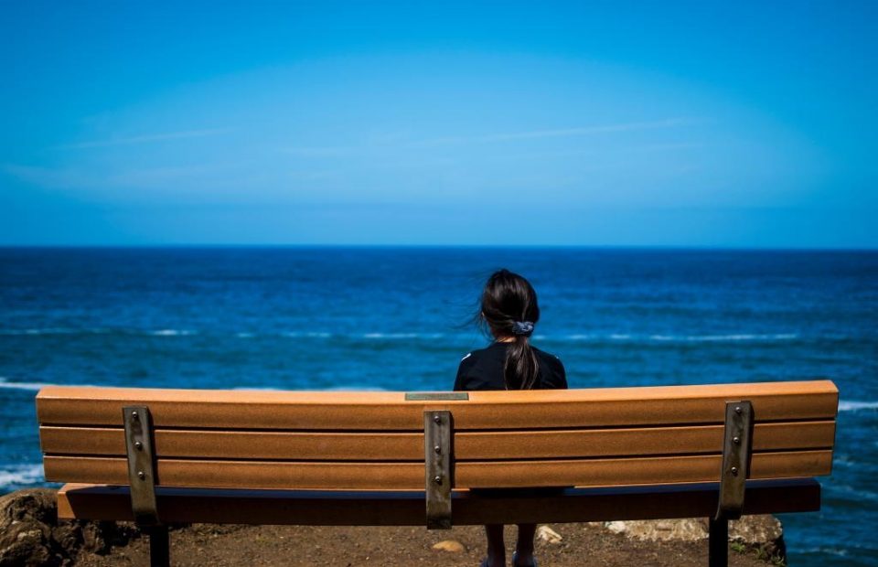 A girl with dark hair tied back sits on a wooden bench overlooking the ocean under a clear blue sky.