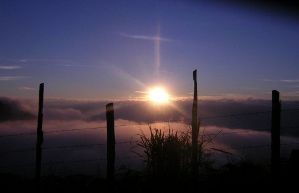 A sunrise or sunset over a cloud-filled sky with a fence in the foreground, and the sun casting a glow with lens flare.
