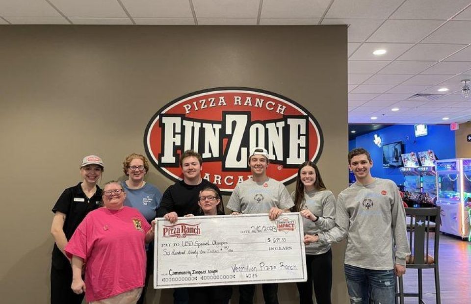 Group of seven people standing in front of a Pizza Ranch FunZone sign, holding a large check in a game arcade area.