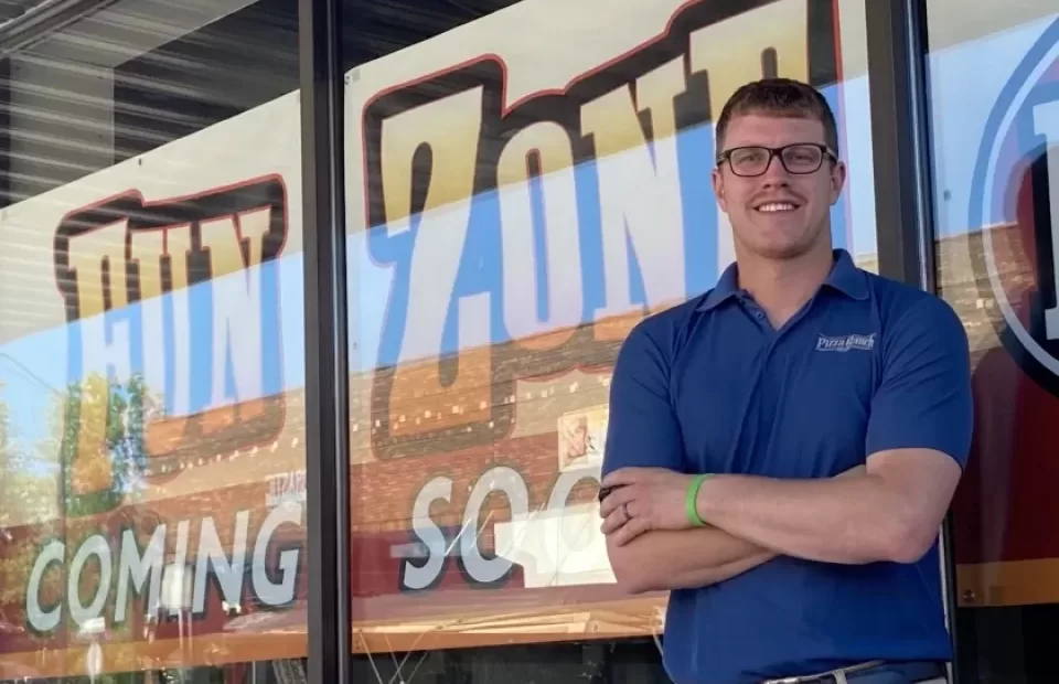 A man in a blue Pizza Ranch shirt and glasses stands outside a building with large windows that display "COMING SOON" signage.