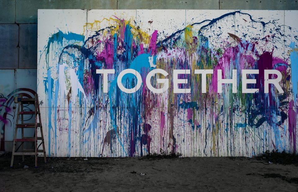 Colorful painted mural with the word "TOGETHER" in white letters, featuring abstract splashes of pink, blue, purple, yellow, and green, on a white background.