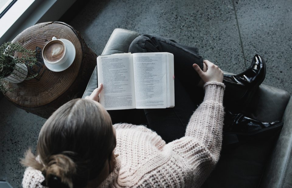 A person with blonde hair, wearing a beige knit sweater and shiny black boots, sitting on a gray armchair, holding an open book in their lap, next to a round wooden side table with a potted plant and a cup of coffee.