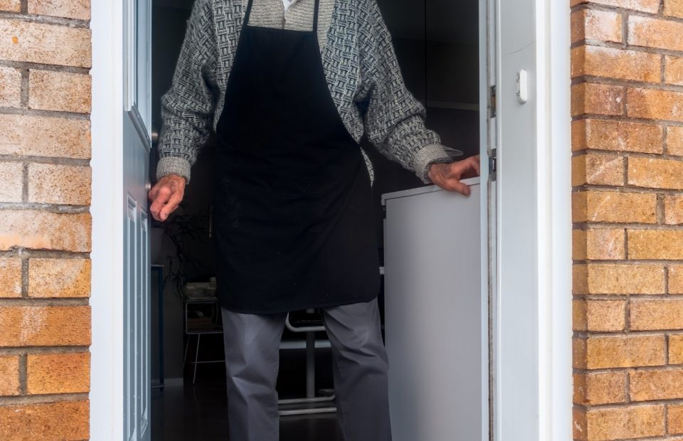 An elderly man wearing glasses, a patterned sweater, and a black apron stands at a doorway, smiling. A blue gift bag and a mug are on the doorstep.