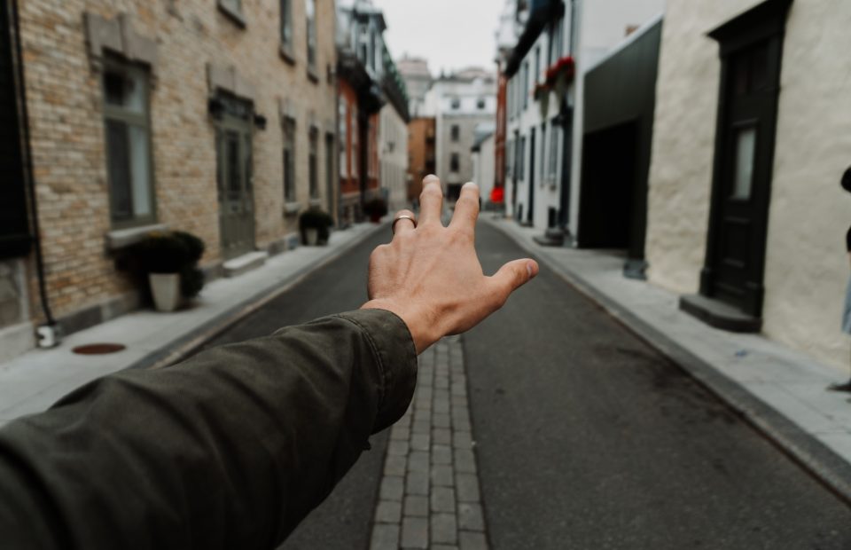 A hand with a ring on the finger reaching out towards a narrow, paved street lined with brick and plaster buildings on both sides.