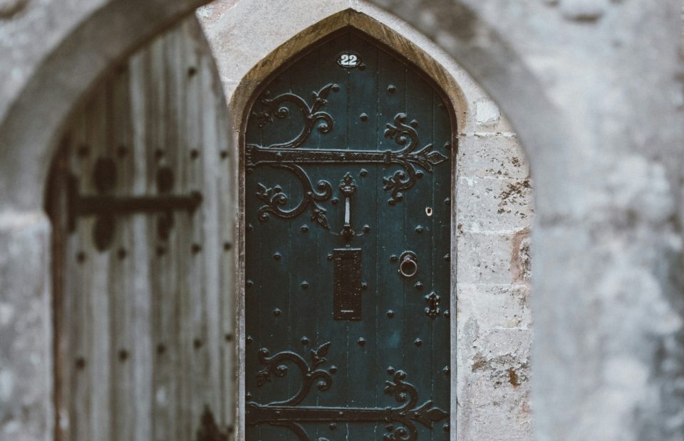 A weathered stone archway with a wooden gate leads to a black metal door with ornate scrollwork and the number 22, framed by aged stone walls.