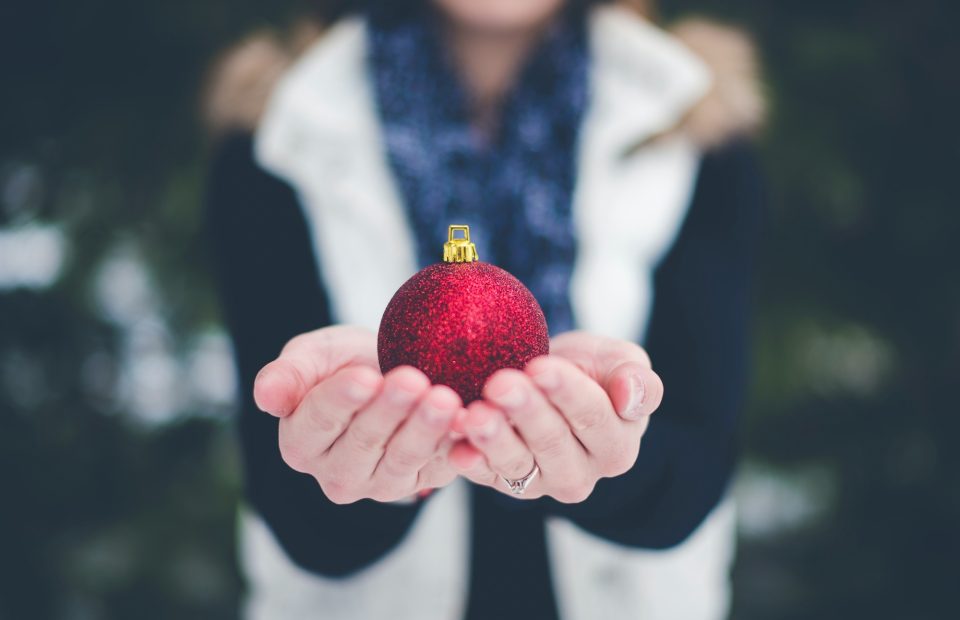 Person holding a red, glittery Christmas ornament with both hands, wearing a dark coat with a white vest and a blue scarf, with blurred greenery in the background.