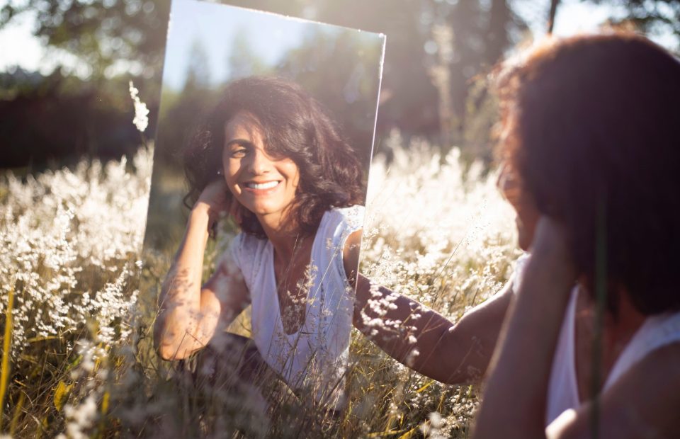 A woman holding a mirror outdoors in a field of white flowers, smiling as she looks at her reflection, with sunlight and trees in the background.
