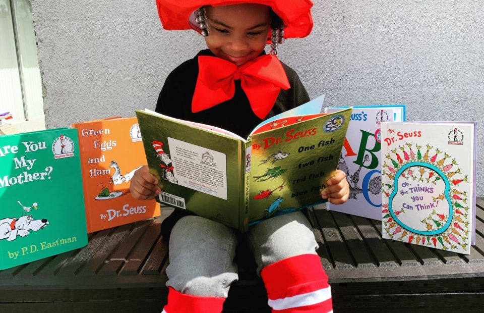 A girl dressed in a holiday-themed outfit with a red and white hat, red bow tie, and striped leg warmers, sitting on a bench and reading a book among other colorful books.