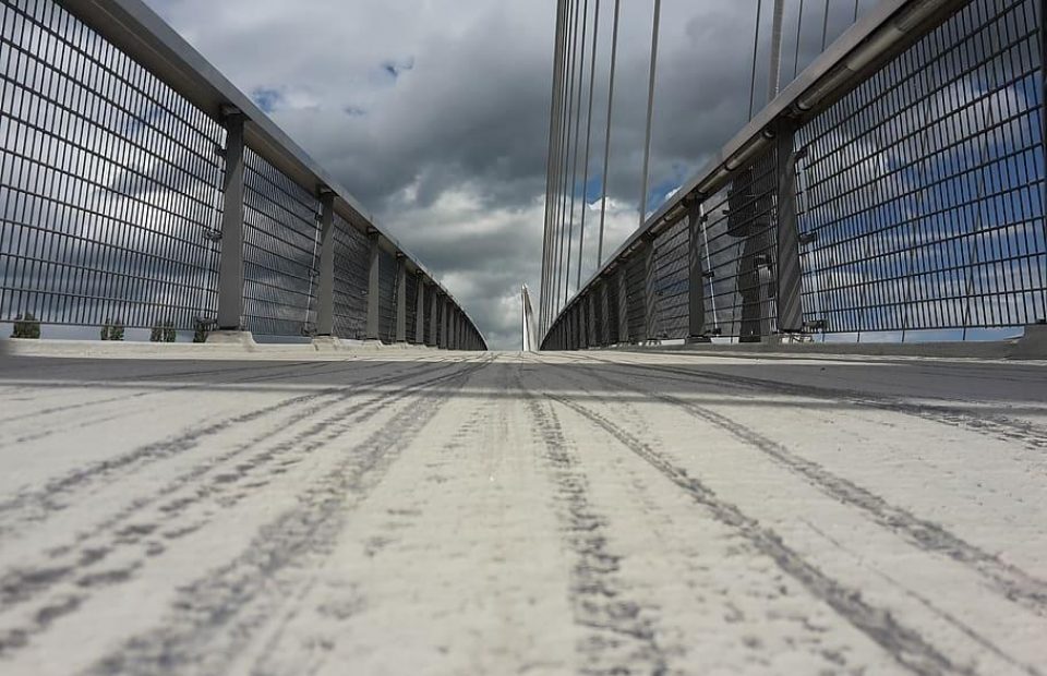 Low-angle view of a bridge with metal railings and cables, cloudy sky overhead, seen from the textured walkway surface. Two figures in uniforms are visible on the right side.