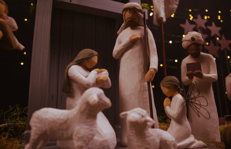 Wooden nativity scene figurines including Mary, Joseph, baby Jesus, and one angel, with a rustic background and soft, warm lighting.