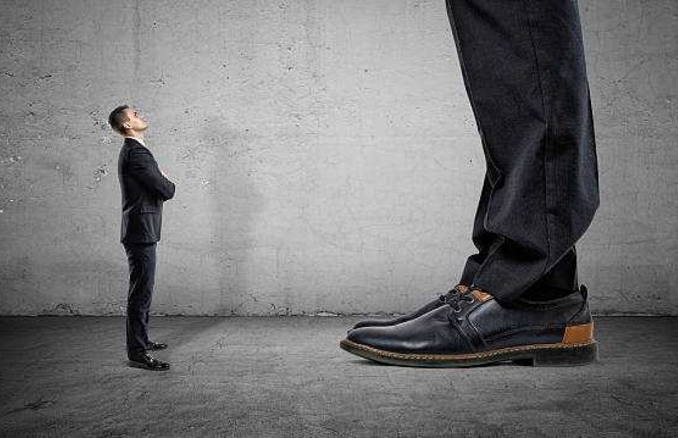 A tiny businessperson in a suit stands in front of an enormous pair of feet in dress shoes, both facing each other against a plain gray wall.