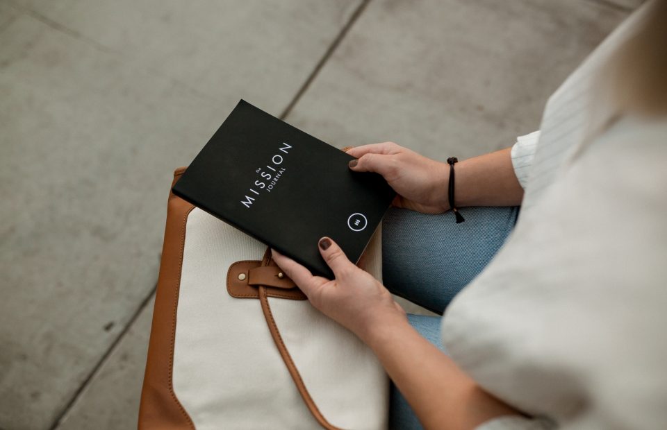A person sitting with a black journal titled "The Mission Journal" in their hands, wearing a white top and a black bracelet, with a tan and cream bag beside them.