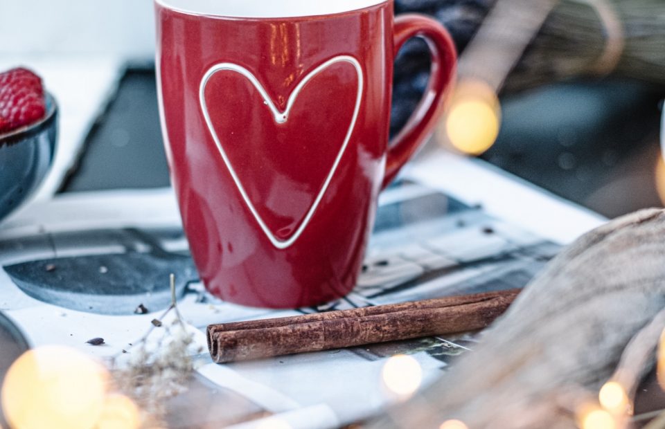 A red mug with a white heart outline sits on a table surrounded by cinnamon sticks, fairy lights, a napkin, and berries in the background.
